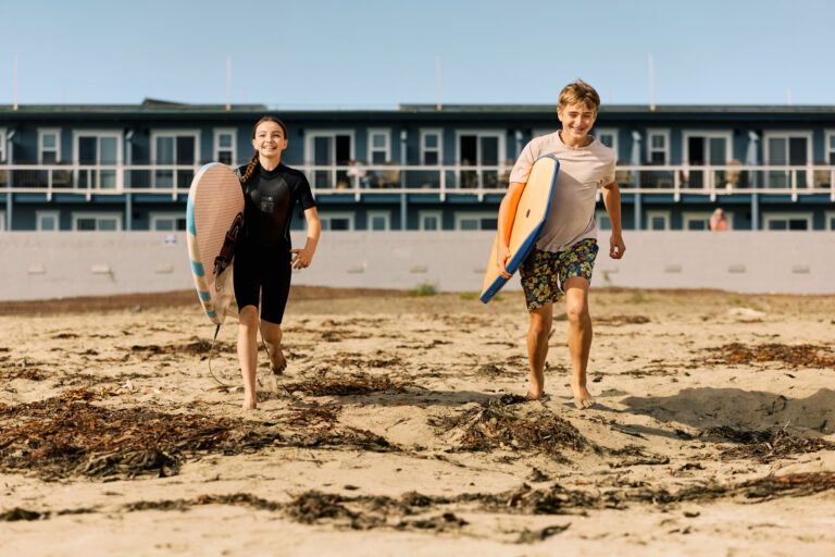 Kids with surfboards running down the beach with hotel in background